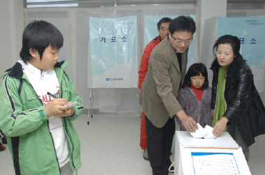 ▲ 정평초교 전교어린이회장인 정재헌(왼쪽) 군이 아빠 엄마가 기표한 투표용지를 투표함에 넣는 모습을 지켜보고 있다.