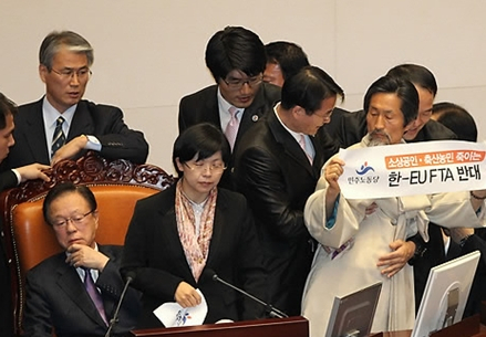되찾은 국회의장석 민노당 강기갑의원이 한EU FTA 반대를 적은 손 플래카드를 들고 있는 가운데, 박희태 국회의장이 잠시 물리적 충돌을 빚었던 국회의장석을 되찾았다.