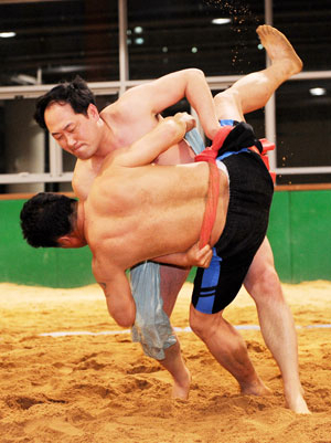 기자가 배지기 기술을 배워 연습상대를 쓰러뜨리고 있다.