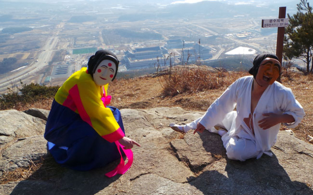 문수지맥이라는 팻말이 박힌 검무산 정상에서 하회별신굿탈놀이 부네탈(손상락)과 이매탈(권오중)이 태극바위를 가리키고 있다. 산 정상 평평한 너럭바위에는 붉은빛이 나는 바위와 푸른빛이 도는 바위가 서로 역S자 모양의 바위틈으로 갈라져 태극무늬를 이루고 있다. 산 아래에 경북도청 신청사가 보인다.