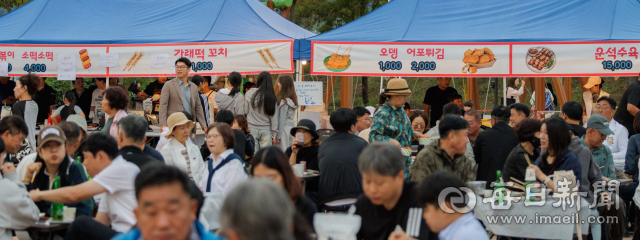 축제장 먹거리 부스 앞에서 방문객들이 음식을 먹으며 이야기를 나누고 있다. 메뉴판에는 